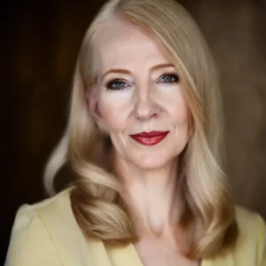 A woman with long blonde hair, blue eyes, and red lipstick, wearing a light yellow top, poses in front of a blurred brown background, looking confidently at the camera.